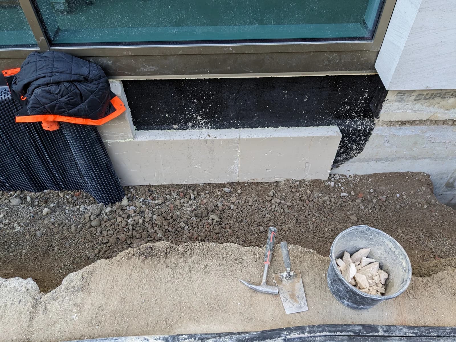 Åpen grøft ved husvegg med synlig svart fuktsikring, hvite isolasjonsplater, knasteplast, spade og hammer.