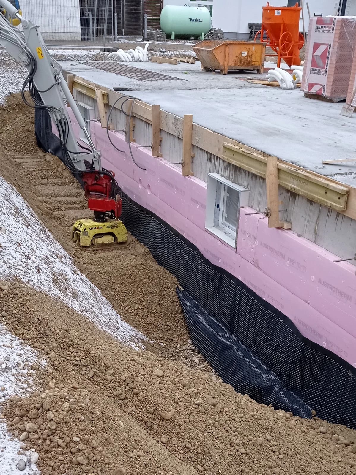 Kjellervegg med rosa utvendig isolasjon, svart knasteplast og gravemaskin som komprimerer fyllmasser langs huset.
