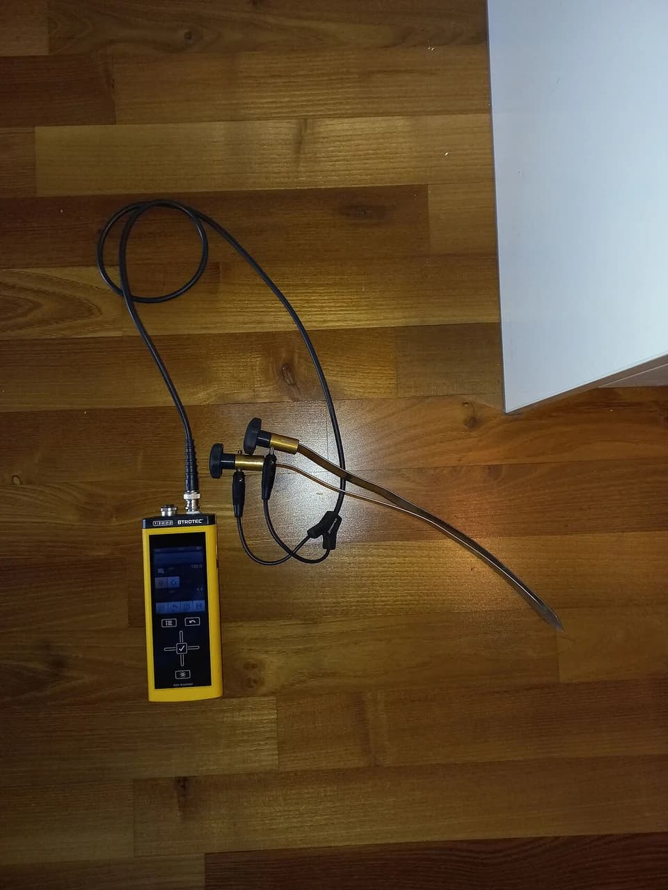 En fuktmåler med tilkoblede sonder ligger på et tregulv ved en vegg.